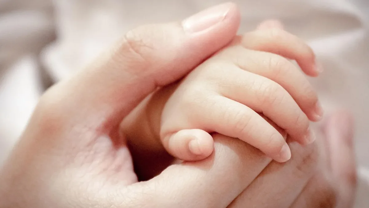A close-up of an adult's left hand gently holding a baby's right hand, with the adult's thumb on top of the baby's thumb and the adult's fin
