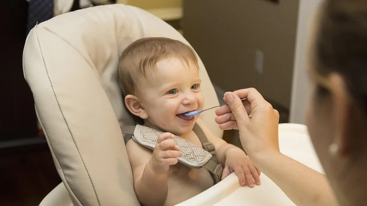 A baby sitting in a high chair, being fed by an adult.